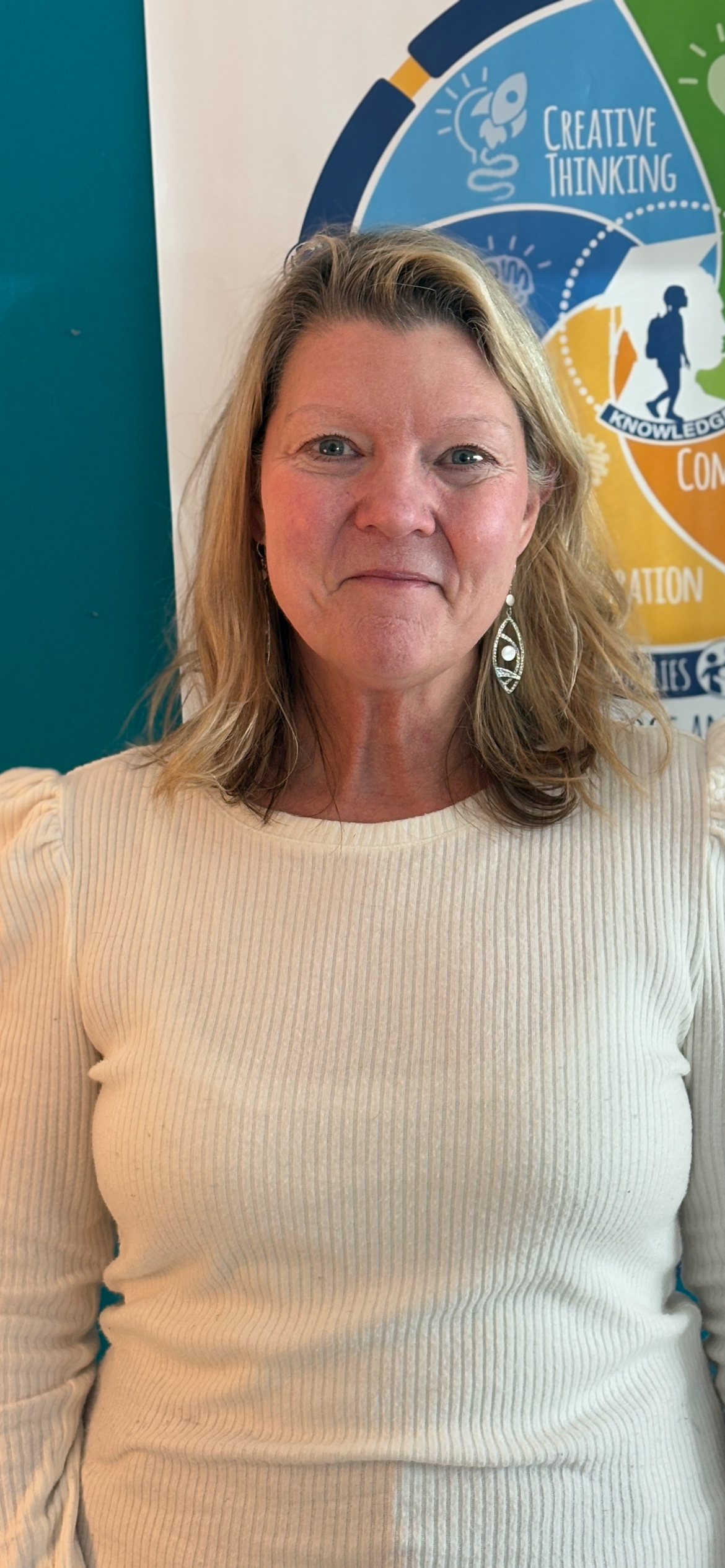 A woman with shoulder-length blonde hair smiles slightly while standing in front of a colorful educational poster. She is wearing a cream-colored, ribbed long-sleeve top and dangling earrings.