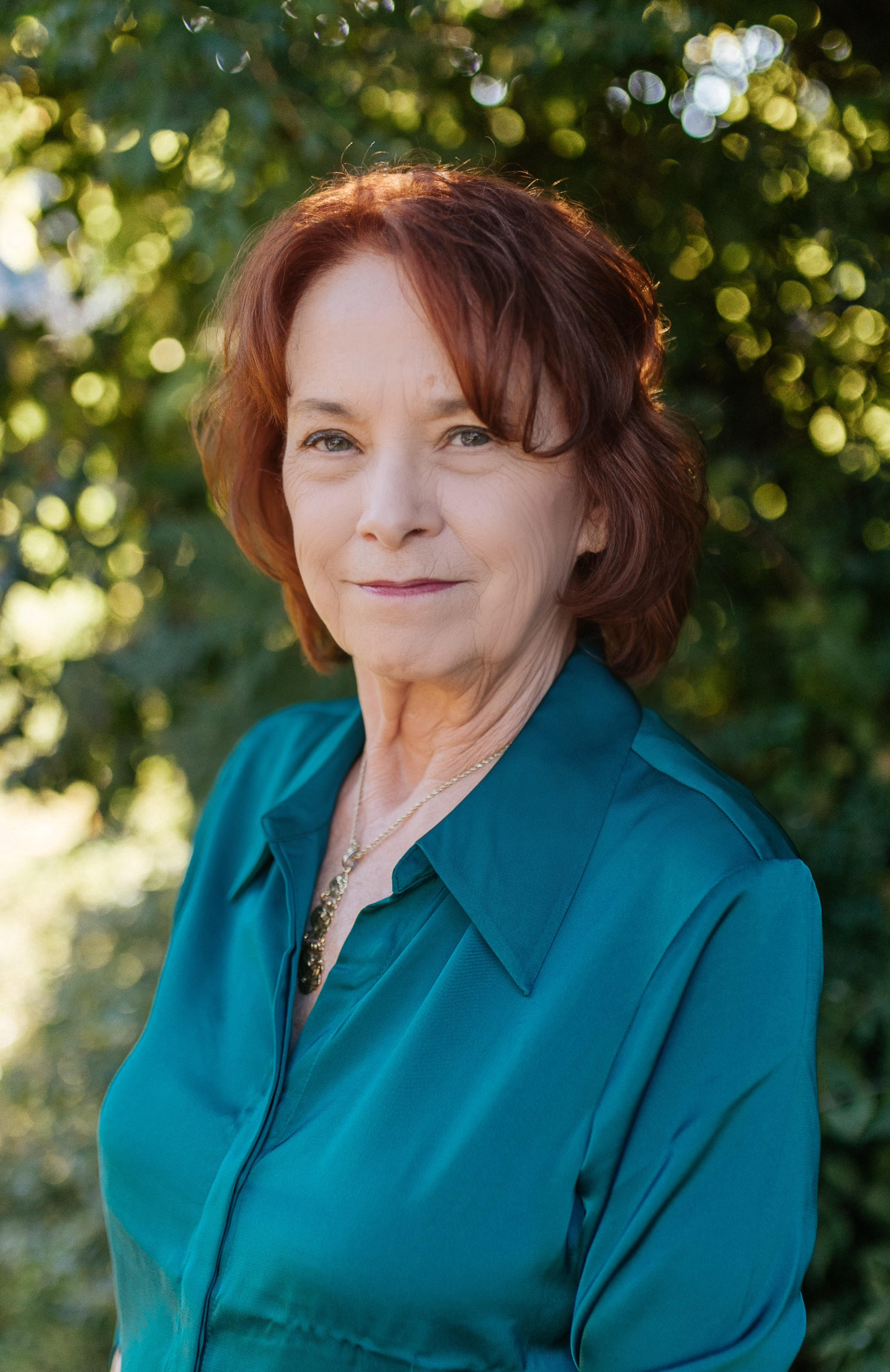 A portrait of a woman with red hair wearing a teal blouse, standing outdoors with sunlight filtering through trees in the background.