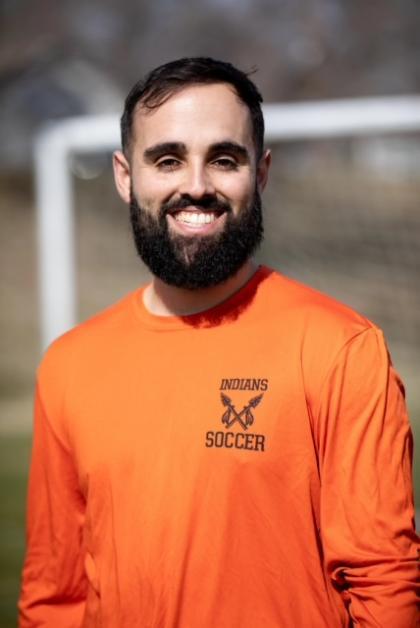 A man smiling outdoors on a sunny day, wearing an orange long-sleeve shirt with the words 